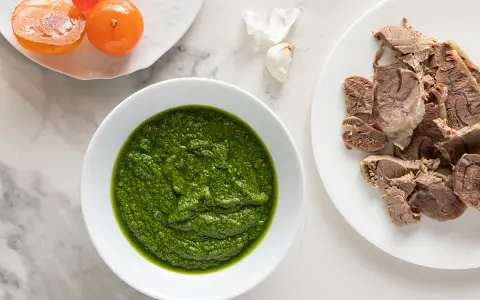 Una ciotola di vibrante salsa verde accanto a fette di carne bollita e pomodorini confit.