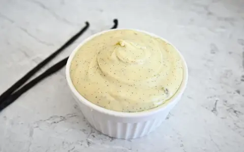 Un bol de ganache chocolat blanc onctueuse, parsemée de points de vanille, avec deux gousses de vanille à côté.
