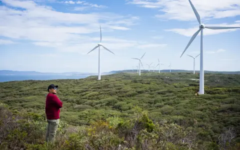 Mann steht vor Windrädern, die die australien energieversorgung repräsentieren. Die Landschaft ist grün und hügelig, mit dem Meer im Hintergrund.