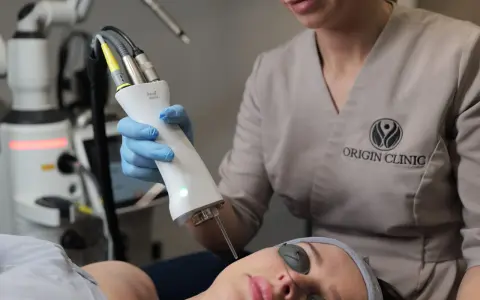 Zabieg laserem Fotona R35NX w Origin Clinic. Widoczna głowica lasera i pacjentka w okularach ochronnych. Oczekiwane spektakularne efekty odmładzające.