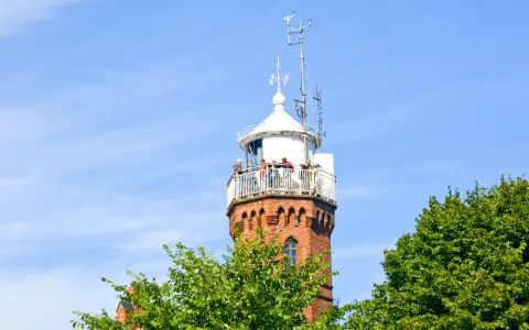 Widok na ceglaną latarnię morską w Ustce z tarasem widokowym, na którym stoją ludzie. Wokół zielone drzewa i błękitne niebo.
