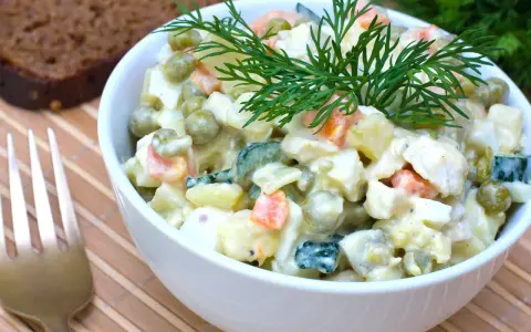 Ein Schüsselchen Olivier Salat, garniert mit Dill, neben einer Scheibe dunklem Brot und einer Gabel.