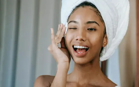 Une jeune femme avec une serviette sur la tête, appliquant un soin sur son visage. Elle semble heureuse, peut-être grâce aux bienfaits du collagène ou de l'acide hyaluronique.