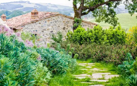 Un chemin de dalles de pierre serpente à travers un jardin luxuriant, bordé de divers types de haies. Des fleurs roses et des arbustes verdoyants entourent une maison en pierre toscane.