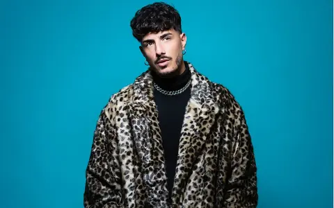 Wade DJ, con su abrigo de leopardo y cadena plateada, posa con confianza ante un fondo azul vibrante.
