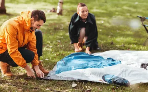 Ein Mann in einem orangen Anorak und eine Frau im Hintergrund bauen ein Zelt auf. Sie bereiten sich auf das **Zelt reinigen** nach dem Camping vor.