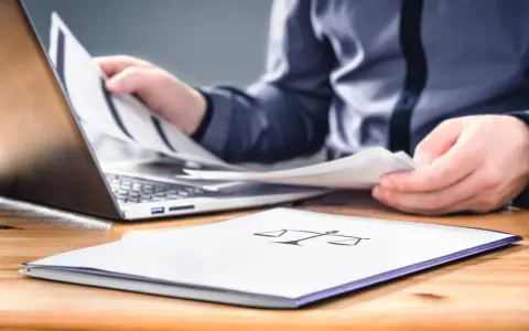 Osoba przegląda dokumenty przy laptopie. Na pierwszym planie folder z symbolem wagi, sugerujący sprawy związane z prawem i potencjalną masą upadłościową.
