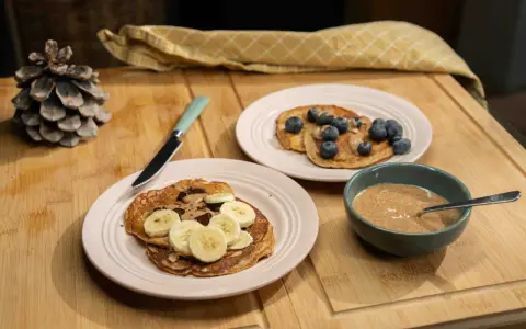 Pancakes ig bas farine complète, garnis de bananes, de chocolat et de myrtilles, avec un bol de beurre d'amande.