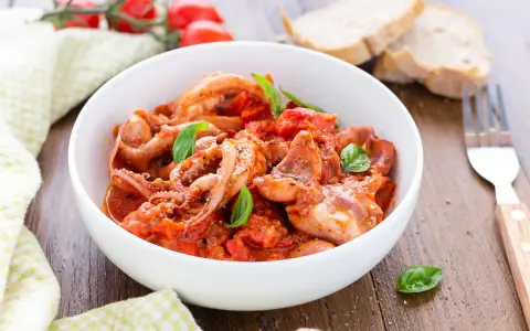 Totani in umido con pomodoro e basilico, serviti in una ciotola bianca, con pane e forchetta sullo sfondo.