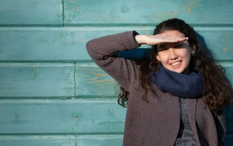 Ragazza sorride, coprendosi gli occhi dal sole. Il fastidio alla luce non le impedisce di godersi la giornata.