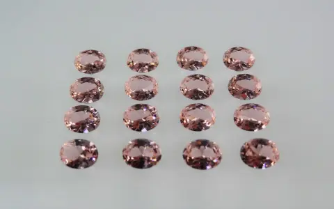 Sedici pietre di morganite ovali, rosa pesca, disposte in file ordinate. Le loro sfaccettature riflettono la luce, mostrando le belle morganite proprietà.