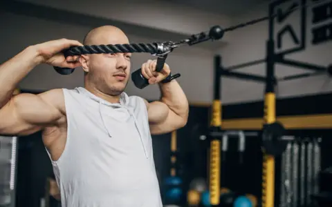 Man med rakad skalle utför face pulls med ett rep i ett gym.
