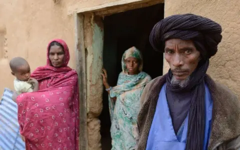 Ein Tuareg-Mann mit schwarzem Turban und ein Kind mit seiner Mutter vor einem Lehmhaus in der Sahara.