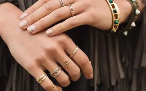 Mani adornate con anelli, tra cui uno con numeri romani, un bracciale colorato e un anello a forma di "S". Non c'è un anello da single specifico, ma la varietà suggerisce stile personale.