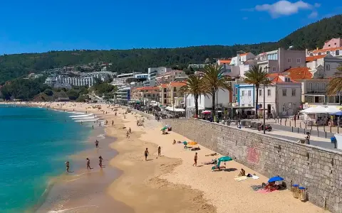 Sonne, Strand und Meer in Sesimbra, Portugal. Menschen genießen den Tag am goldenen Sandstrand, während im Hintergrund die Stadt und grüne Hügel aufragen.