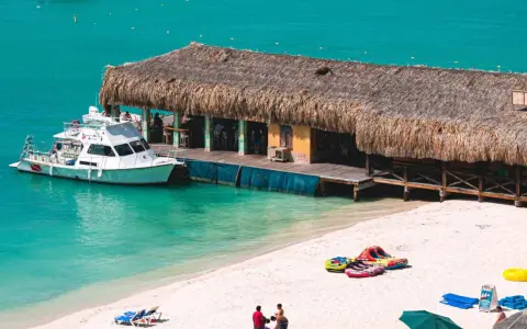 A boat docks at a thatched-roof beach bar on a sunny day. This scene embodies the welcoming spirit of Aruba, a destination known for being gay friendly.