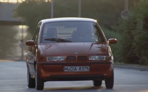 Pomarańczowy BMW E1 jedzie po drodze, światła odbijają się od karoserii.