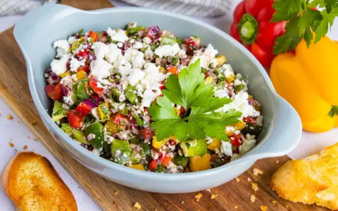 Bunter Paprikasalat mit Feta, garniert mit Petersilie, serviert in einer hellblauen Schüssel auf einem Holzbrett. Daneben liegen geröstete Brotscheiben.