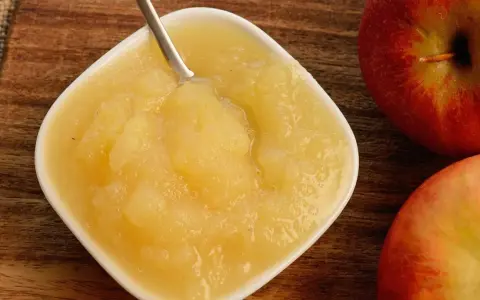 Frisches Apfelmus mit Schale, zubereitet im Thermomix, in einer weißen Schale auf Holzuntergrund. Daneben liegen zwei rote Äpfel.