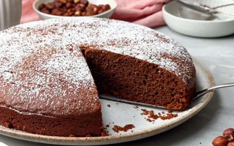 Ein saftiger Nutella Kuchen, bestäubt mit Puderzucker, wartet darauf, angeschnitten zu werden. Daneben eine Schüssel mit Haselnüssen.