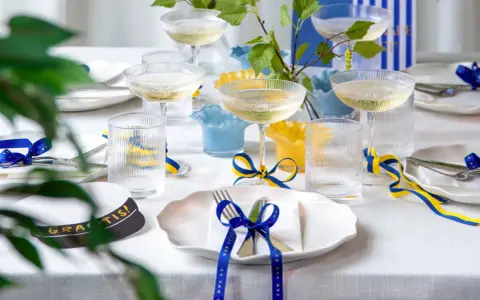 Festligt dukat bord med champagneglas, blå och gula band som knyts i en blå rosett, samt en studentmössa.