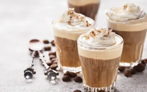 Tre bicchieri di deliziosa crema al caffè, guarniti con panna montata e cacao. Chicchi di caffè e cucchiaini decorati completano la scena.