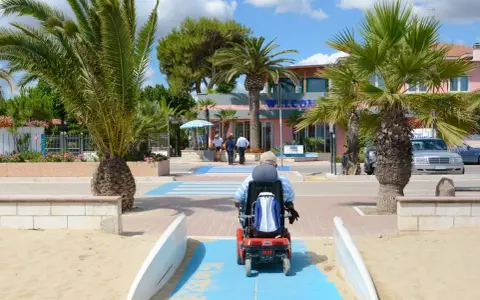 Accesso facilitato per tutti nei villaggi in Abruzzo sul mare. Un uomo in carrozzina elettrica percorre una passerella blu sulla sabbia, tra palme rigogliose e un edificio con la scritta "Welcome".