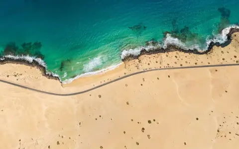 Luftaufnahme eines einsamen **Surferstrands** auf **Fuerteventura**. Türkisblaues Meer trifft auf goldgelben Sand und eine felsige Küste. Eine Straße schlängelt sich entlang der Küste.