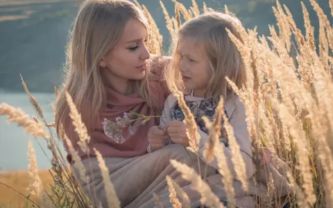 Matka i córka w trawach. Córka z autyzmem skupiona na kwiatku, matka z czułością patrzy na nią.