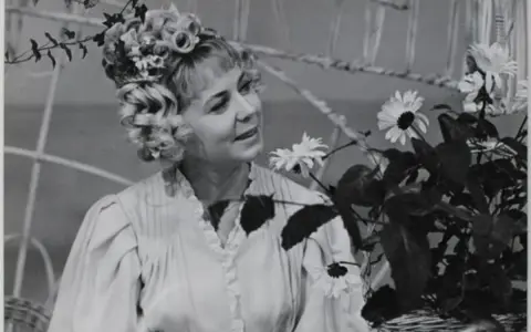 Ilona Stawińska w kwiecistej fryzurze i jasnej sukni, otoczona kwiatami, wygląda jak z baśni.