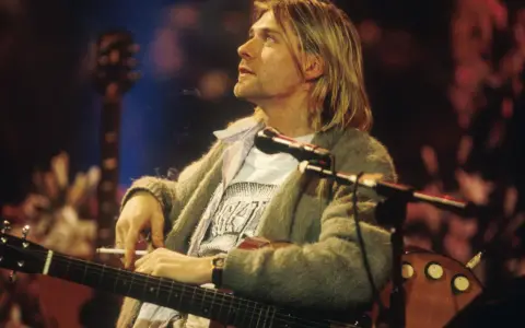 Kurt Cobain z papierosem w dłoni, grający na gitarze. Jego muzyka to część 100 najlepszych piosenek wszechczasów.