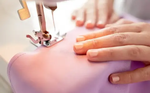 Dłonie prowadzą różowy materiał pod stopkę maszyny do szycia. Widać igłę i pedał.