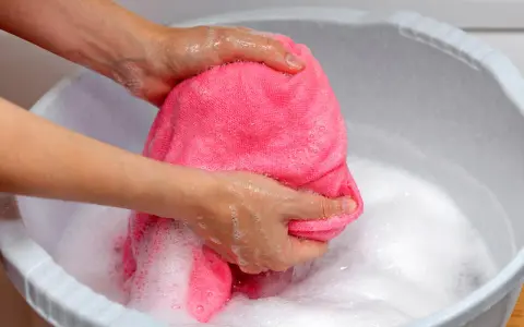 Delikatne pranie wełny merino w pianie. Ręce ugniatają różowy materiał w misce z wodą.