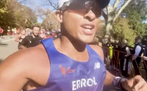 En löpare i blått linne, med texten "ERROL", kämpar på för att springa marathon. Publiken hejar längs banan.