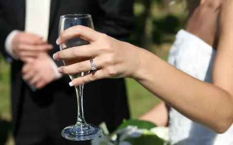 Dłoń panny młodej z pierścionkiem, wznosząca kieliszek szampana. W tle pan młody. 34 rocznica ślubu nazwa.