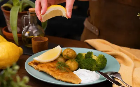 En hand pressar citron över en tallrik med panerad torsk, potatis, broccoli och en klick sås.
