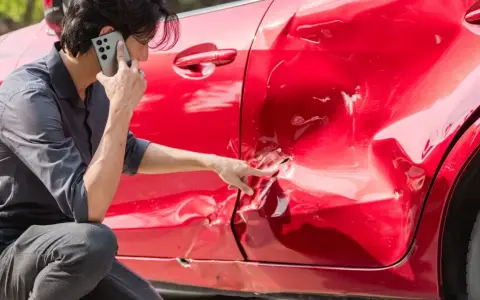 Hombre llama por teléfono señalando el grave golpe en la puerta de su coche rojo.