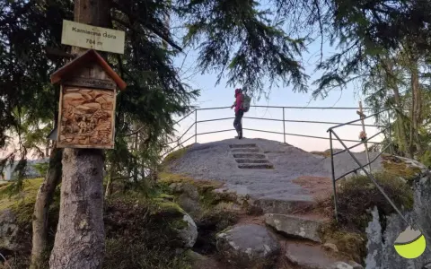 Widok na szczyt Kamienna Góra (704 m). Turystka podziwia panoramę, a obok drzewa ozdobiona kapliczka.