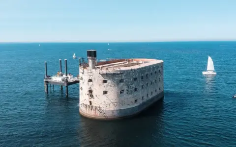 Fort Boyard, potężna twierdza na morzu, otoczona błękitną wodą. Obok platforma wiertnicza i żaglówka.