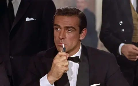 James Bond, elegante in smoking, accende una sigaretta con un'aria pensierosa.