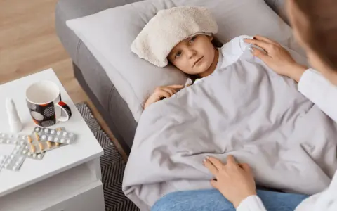 Chora dziewczynka z okładem na głowie leży pod kocem. Obok leki i kubek. Mama czuwa, zapewniając opiekę na dziecko.