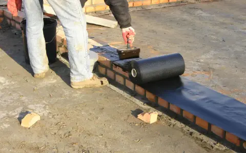 Pracownik nakłada masę bitumiczną na ceglaną ścianę, przygotowując ją do położenia hydroizolacji z rolki.
