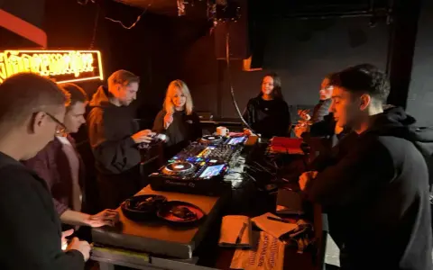Grupa młodych ludzi przy konsoli DJ-skiej, uczących się, jak zostać DJ-em. Neonowy napis w tle.
