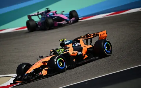 Un coche de Fórmula 1 naranja y negro compite en la pista, anticipando el reglamento F1 2026. Otro coche rosa y azul le sigue de cerca.
