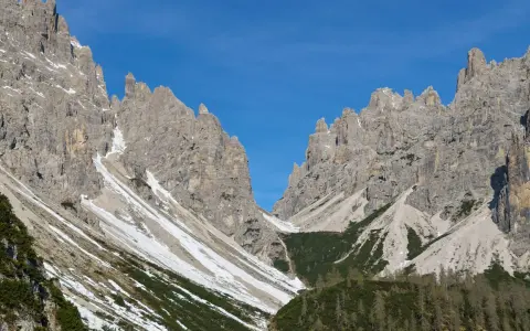 Vette rocciose imponenti sotto un cielo azzurro. Un sentiero scende verso il rifugio Giaf, tra pini e chiazze di neve.