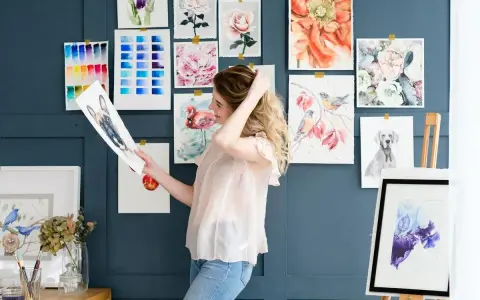 Mujer joven sonríe mientras revisa un dibujo de perro, rodeada de acuarelas de flores, aves y un flamenco. Un espacio inspirador para estudiar dibujo.