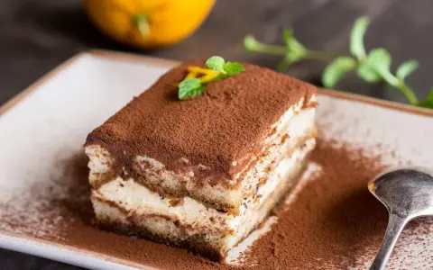 Pyszne tiramisu na biszkopcie, posypane kakao, z miętą i skórką pomarańczy. Obok łyżeczka i pomarańcza.