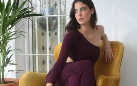 Mujer con escotes asimétricos en un sillón amarillo, junto a una planta y una ventana con vidrieras.