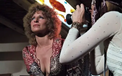 Barbra Streisand poses as Audrey Hepburn, dressed in a sparkly outfit, captures the moment with her camera.