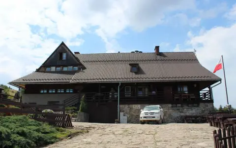 Drewniana bacówka Biała Woda z białym SUV-em na kamienistym podjeździe. Biało-czerwona flaga powiewa na maszcie.